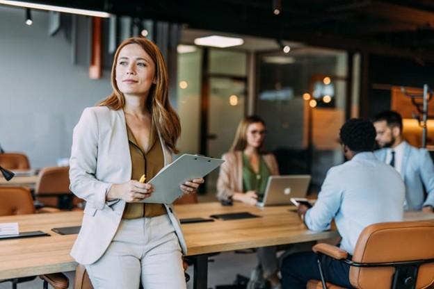 A business executive standing in an office while co-workers meet in the background.