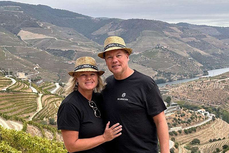 Photo of a smiling couple in straw hats overlooking terraced vineyards and a river.