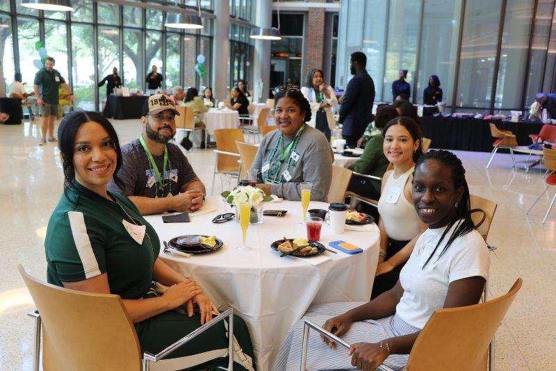 Five diverse people smile around a table with drinks and food at an indoor event.