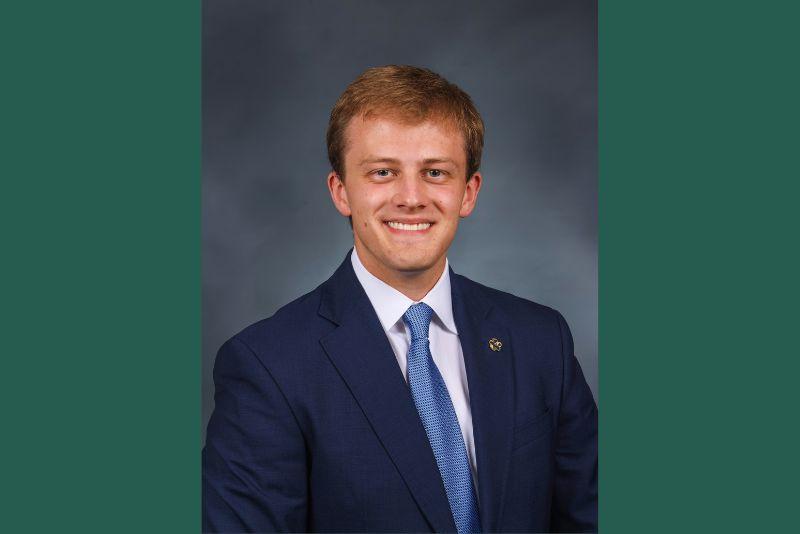 Smiling young man in a blue suit, white shirt, and blue tie against a dark background.