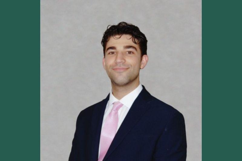 A professional headshot of a smiling young man in a dark suit and pink tie against a gray background. 