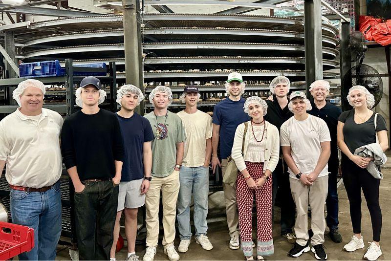 Group of people in hairnets posing in a bakery with a large spiral conveyor.
