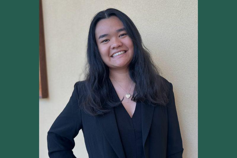 Smiling woman with long dark hair, wearing a black blazer and heart necklace.