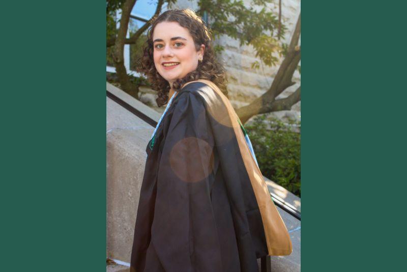 Smiling graduate in black gown and academic hood on a sunny day.