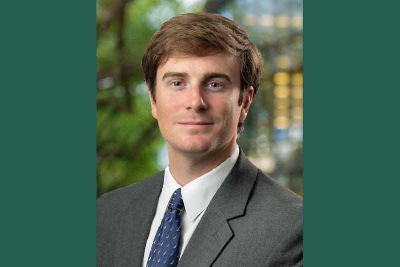 Headshot of a man with light brown hair, gray suit, blue tie. Blurred green background.