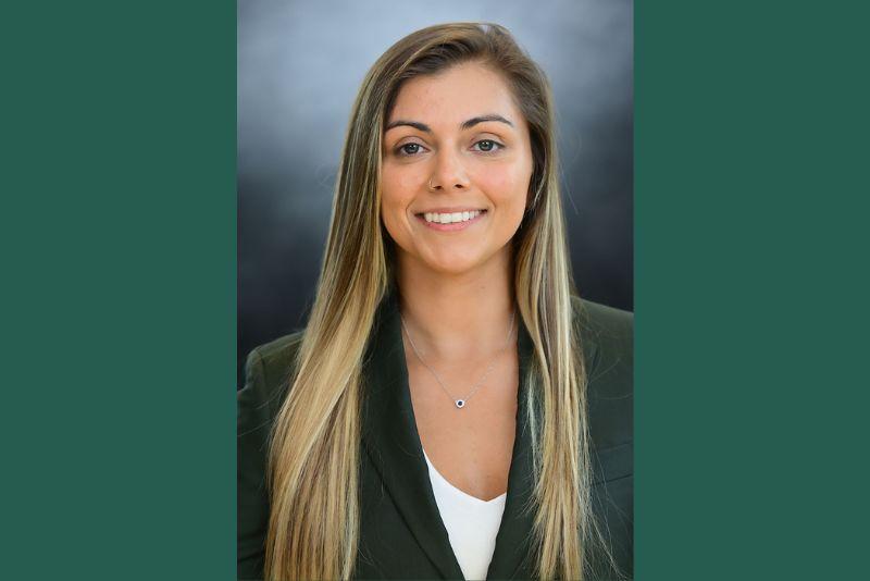 Smiling young woman with long blonde hair and a dark blazer on a gray background.