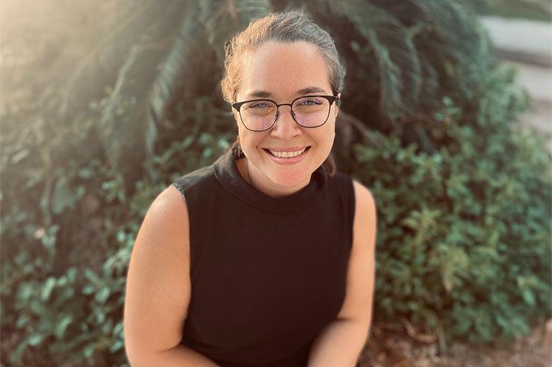 Woman in black shirt and glasses smiles warmly outdoors.