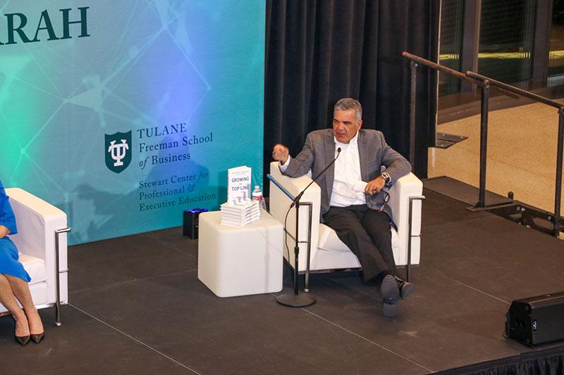 Speaker in grey suit gestures on stage next to another speaker and "Growing Top Line" books.