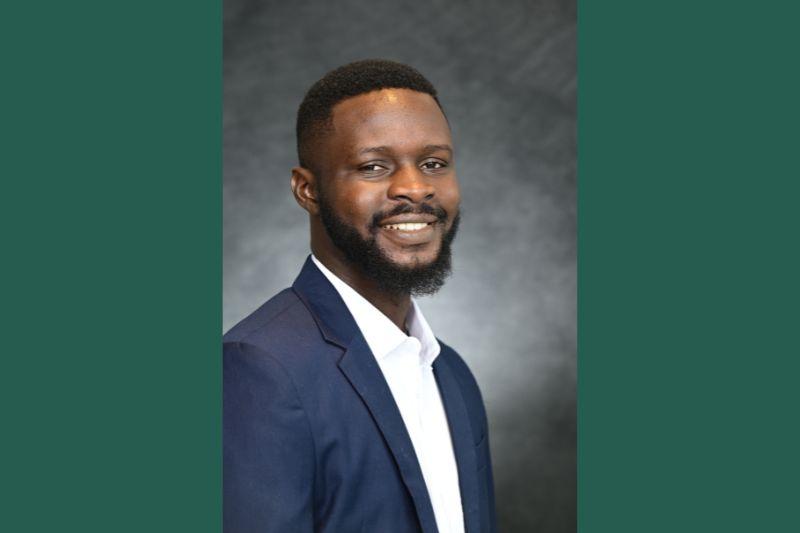 Photograph of a smiling Black man with a beard in a navy suit and white shirt.