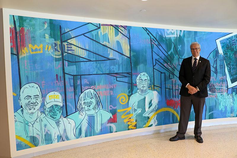 Man in suit standing before a colorful mural featuring graffiti and sketched figures.