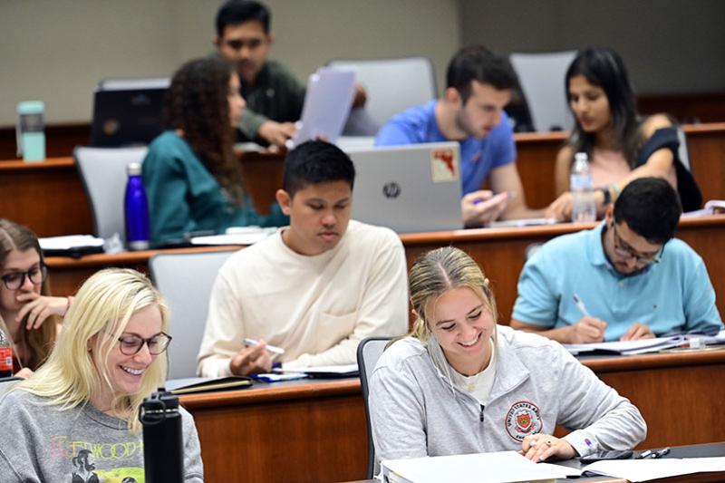 College students smiling and working in a tiered lecture hall classroom.