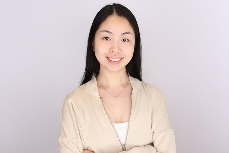 Smiling young woman with dark hair in a beige jacket against a gray background.