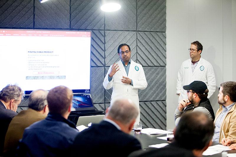 Man in white lab coat presents to an attentive audience in a modern conference room, colleague watches.