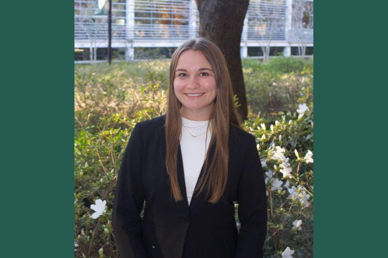 Smiling young woman in a black blazer stands in a garden with white flowers.
