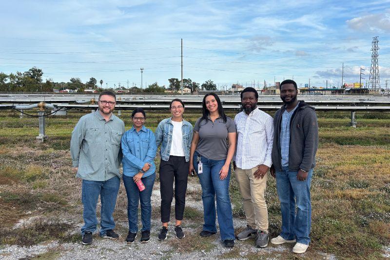 Six smiling people, diverse group, stand in a field with solar panels behind them.