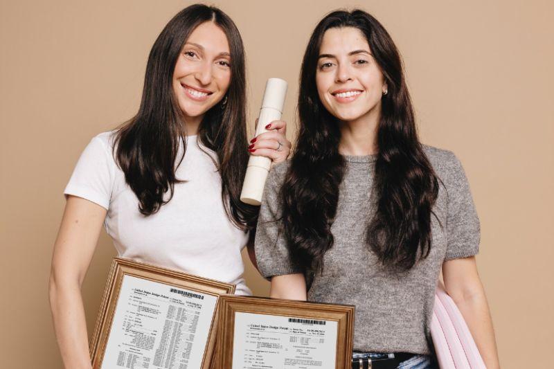 Two smiling women proudly display framed patent certificates and rolled documents.