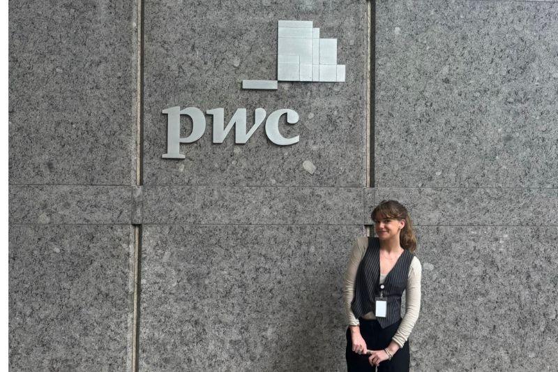 Young woman smiling in front of a gray stone wall with a silver PwC logo.