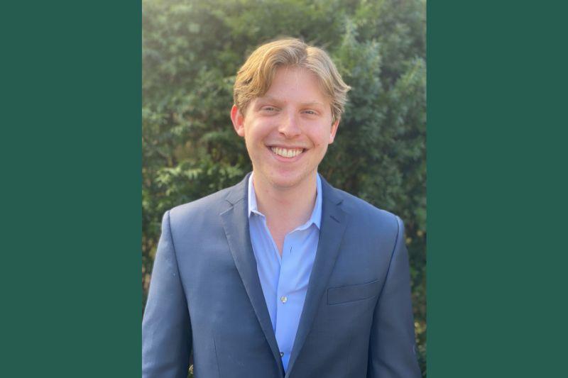 Smiling young man in a dark suit and light blue shirt, green outdoor background.