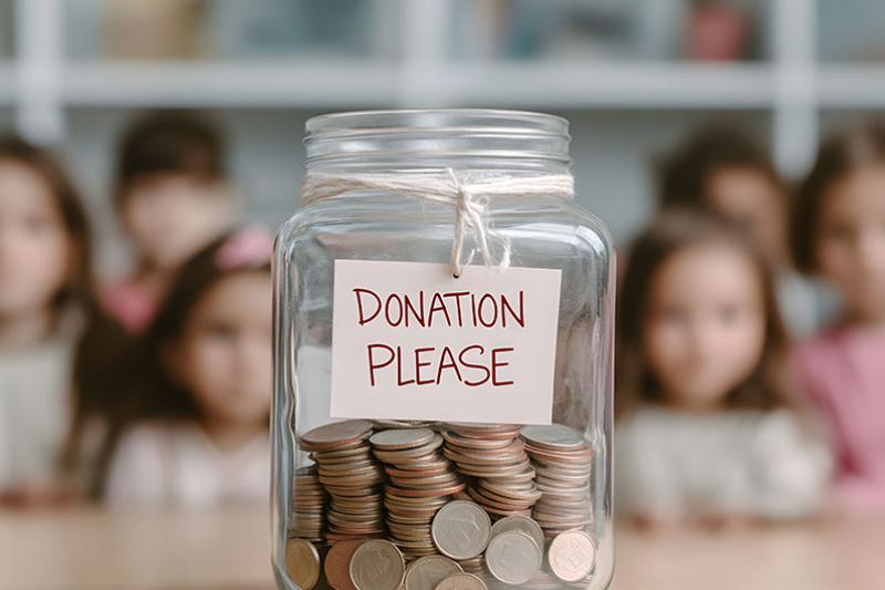 Donation jar filled with coins, labeled "DONATION PLEASE", with blurry children in the background.