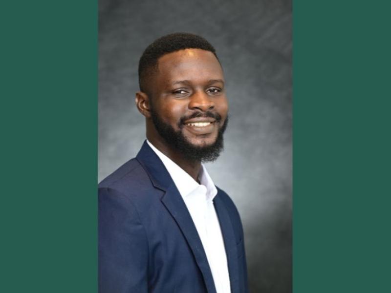 Photograph of a smiling Black man with a beard in a navy suit and white shirt.