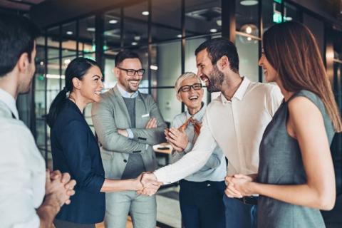 Two business professionals shake hands while coworkers look on.