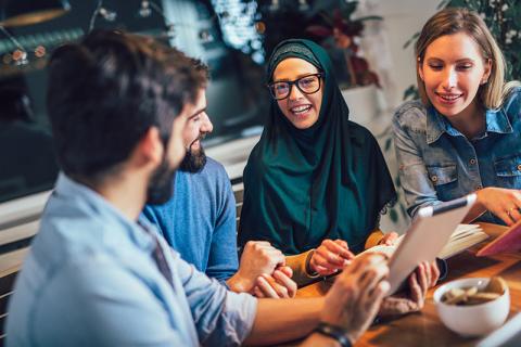 Three smiling people, one in a dark green hijab, looking at a tablet.