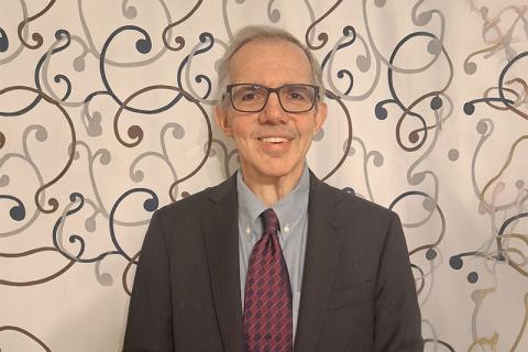 Gus De Franco wearing dark jacket with red tie standing in front of wall