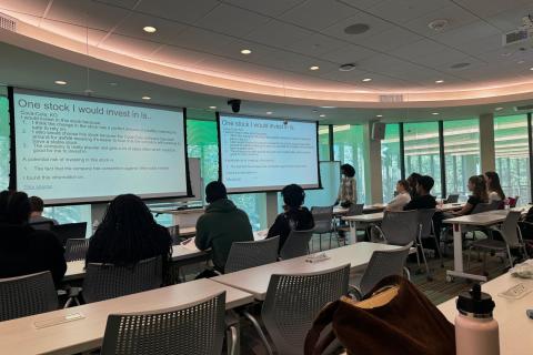 Students in a classroom listen to a presenter standing in front of a screen. 