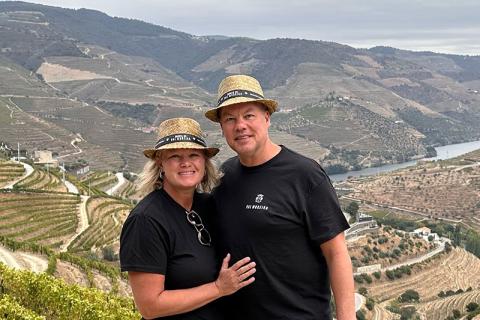 Photo of a smiling couple in straw hats overlooking terraced vineyards and a river.