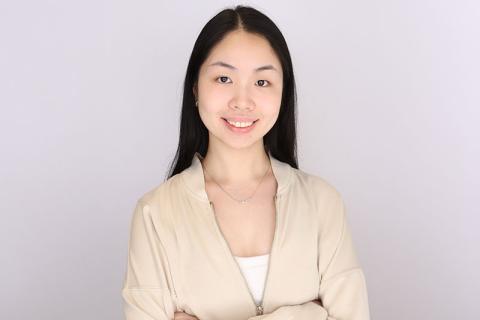 Smiling young woman with dark hair in a beige jacket against a gray background.