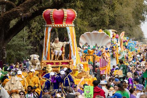 King Rex in a golden costume on a large crown-shaped float at a vibrant Mardi Gras parade.