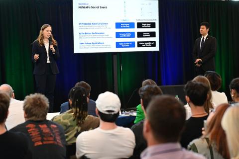 Woman speaks into microphone, man stands by large presentation screen, audience watches.