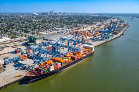 Aerial view of a large container ship docked at a busy port with cranes and a city skyline.