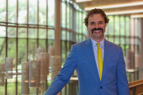 Man with a mustache, curly hair, blue suit, and yellow tie, smiling in an indoor setting.