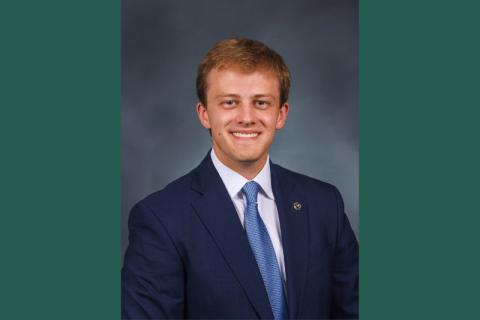 Smiling young man in a blue suit, white shirt, and blue tie against a dark background.