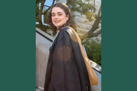 Smiling graduate in black gown and academic hood on a sunny day.