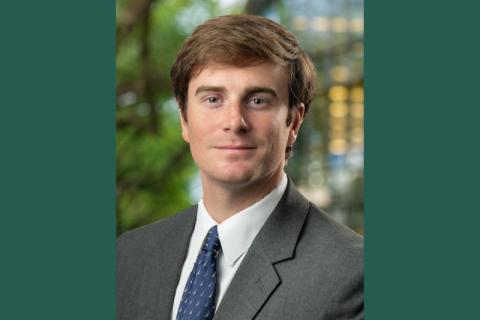 Headshot of a man with light brown hair, gray suit, blue tie. Blurred green background.